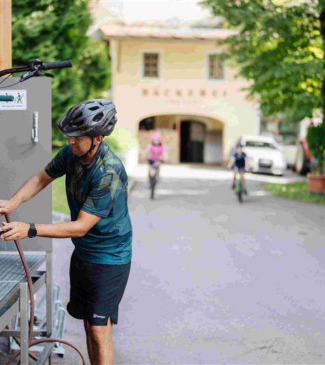 Ein Mann repariert ein Fahrrad in einer malerischen Umgebung. Im Hintergrund fahren weitere Radfahrer und es gibt grüne Bäume sowie Blumen.