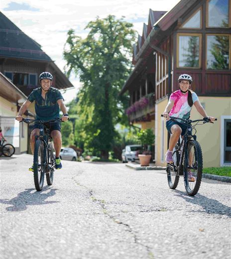 Zwei Personen fahren auf ihren Fahrrädern in einer malerischen Straße. Umgeben von schönen Gebäuden und grüner Natur erleben sie einen sonnigen Tag.